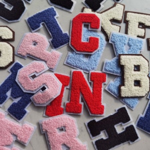 May include: A collection of felt letter patches in various colours, including pink, red, blue, white, and black. The letters are arranged in a random order and include the letters A, B, C, F, H, I, M, N, R, S, and V.