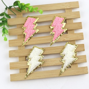 May include: Four lightning bolt shaped iron-on patches. Two are pink with gold glitter trim and two are white with gold glitter trim.