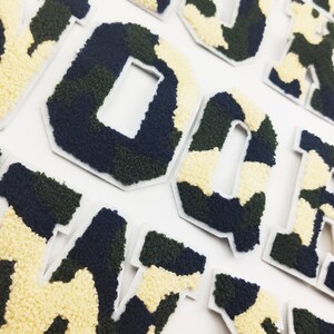 May include: A set of felt letters in a camouflage pattern. The letters are in a variety of colors, including green, black, and white. The letters are all uppercase and are arranged in a random order.