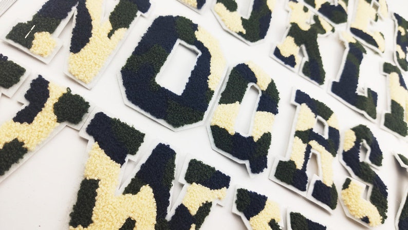 May include: A set of felt letters in a camouflage pattern. The letters are in a variety of colors, including green, black, and white. The letters are all uppercase and are arranged in a random order.