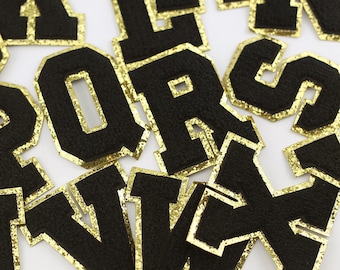 Può includere: Un insieme di lettere in feltro nero con bordino in glitter dorato. Le lettere sono di diverse dimensioni e stili, e sono disposte in ordine casuale.