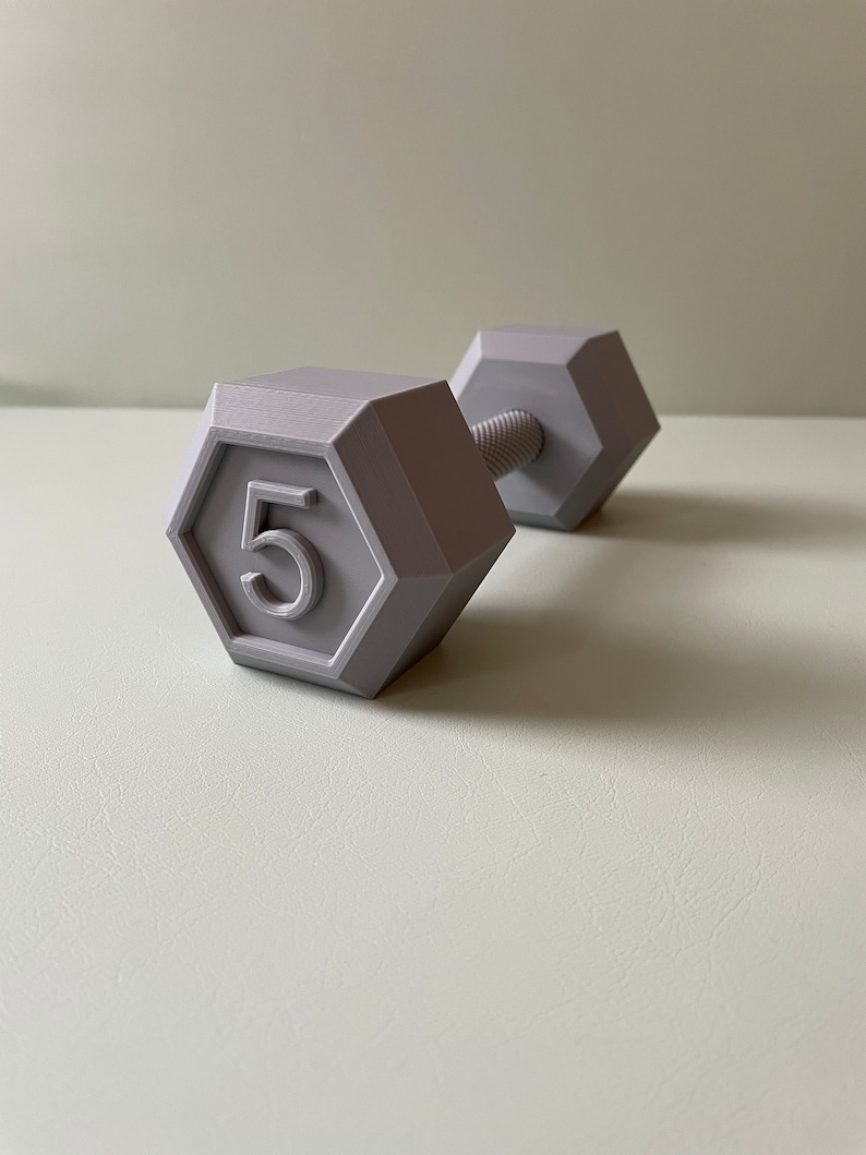 May include: A gray 5-pound dumbbell with a hexagonal shape. The dumbbell is made of plastic and has a smooth surface.