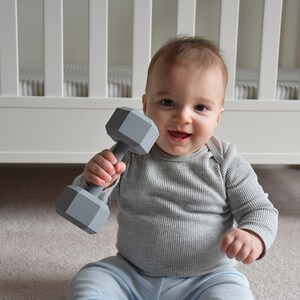 May include: A baby sitting on a gray carpet holding a gray plastic dumbbell toy.
