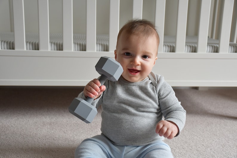 May include: A baby sitting on a gray carpet holding a gray plastic dumbbell toy.