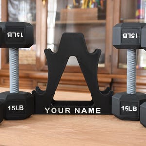 May include: A black dumbbell rack with four black dumbbells. The dumbbells are labeled 10LB and 15LB. The rack has a white label that says "YOUR NAME".