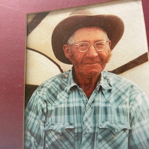 Op de afbeelding: Een zwart-witfoto van een man die een geruit overhemd en een bruine cowboyhoed draagt. Hij glimlacht en kijkt naar de camera. De foto is ingelijst in een rood frame.