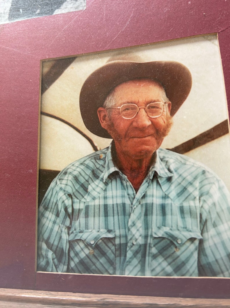 Op de afbeelding: Een zwart-witfoto van een man die een geruit overhemd en een bruine cowboyhoed draagt. Hij glimlacht en kijkt naar de camera. De foto is ingelijst in een rood frame.
