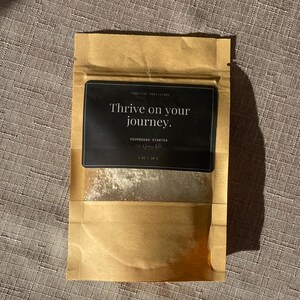 Op de afbeelding: Een bruine papieren zak met een wit label waarop staat "Thrive on your journey."  SOURDOUGH STARTER - 1 OZ / 28 G. De zak bevat een wit poeder.