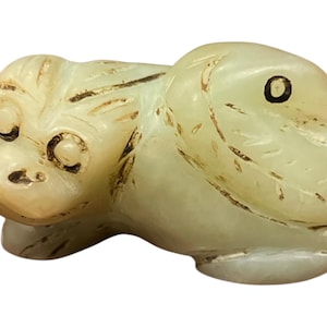 May include: A carved, off-white jade figurine of a stylized animal, possibly a monkey or rabbit. The sculpture has dark brown etched details and black circular eyes. The animal is in a seated position.
