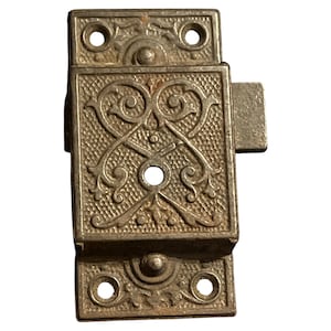 May include: An antique metal cabinet latch with an ornate, symmetrical design. The latch is rectangular with decorative scrollwork and a textured background. It has a small, rectangular catch and is likely made of brass or a similar metal.