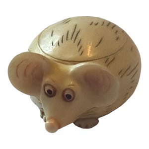 May include: A whimsical, beige-colored mouse figurine with large, rounded ears and a long, pink nose. The mouse has dark eyes and a slightly rounded body, with short, dark lines etched on its back. This decorative item is likely a collectible or a decorative piece.