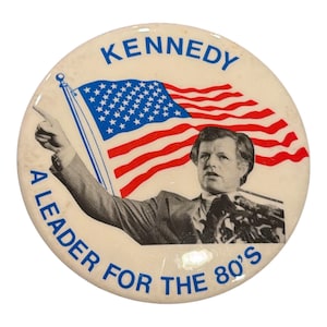 May include: A vintage campaign button featuring a portrait of a man with the text "KENNEDY" and "A LEADER FOR THE 80'S". The button includes an American flag design in red, white, and blue.