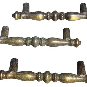 May include: Three antique brass drawer pulls with a decorative design. Each pull features a central, bulbous section flanked by smaller, detailed elements and curved handles. The brass has a weathered, aged appearance.