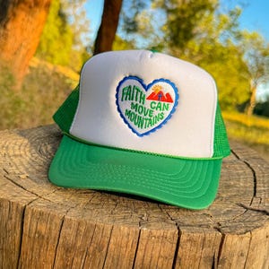 May include: A green and white trucker hat with the text "Faith Can Move Mountains" inside a heart-shaped design. The hat has a green brim and mesh back, and is sitting on a wooden stump.