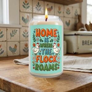 May include: A glass jar candle with a lit wick, featuring a light blue label with the text "HOME IS WHERE THE FLOCK ROAMS" in orange and green letters, surrounded by illustrations of two white chickens, small flowers, and green leaves.