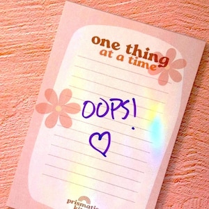 May include: A pink notepad with the text "one thing at a time" and "OOPS!" written on it in purple ink. The notepad has a floral design and the brand name "prismatic kitty" at the bottom.