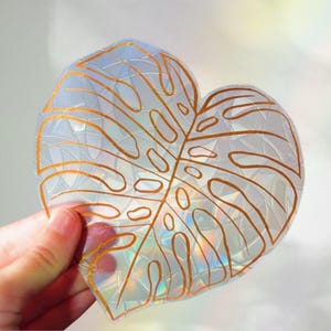 Könnte beinhalten: Ein klares, herzförmiges Dekorationselement mit einem goldfarbenen Umriss eines Monstera-Blattes. Das Element hat eine strukturierte, irisierende Oberfläche, die das Licht reflektiert und einen Regenbogeneffekt erzeugt. Das Design wird von einer Hand gehalten.