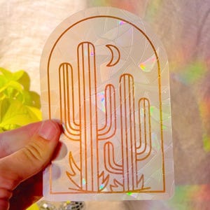 May include: A white iridescent sticker with a gold outline of three cacti and a crescent moon. The sticker has a cracked glass effect.
