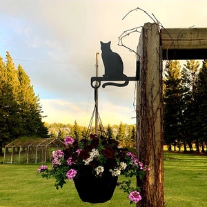 May include: A black metal cat silhouette planter hanger with a hanging basket filled with pink and white flowers. The hanger is attached to a wooden post with vines.