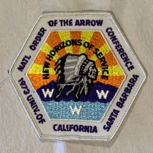 Puede incluir: Un parche hexagonal blanco con costuras moradas y blancas. El parche presenta un tocado nativo americano con un sol radiante amarillo, naranja y rojo detrás. El texto "NATL ORDER OF THE ARROW NEW HORIZONS OF SERVICE CONFERENCE 1973 UNIV. OF CALIFORNIA SANTA BARBARA" está impreso alrededor del borde del parche.