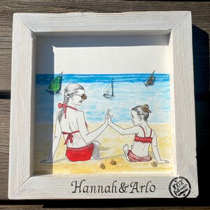 May include: A framed watercolor painting of two people in swimsuits on a beach. The people are holding hands and smiling. The painting is titled "Hannah & Arlo" and has a small, round sticker in the bottom right corner that says "100% handmade with love".
