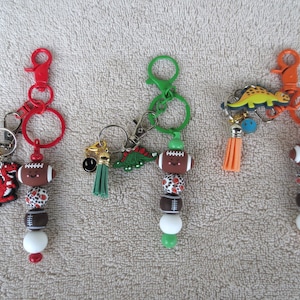 May include: Three keychains featuring colorful plastic clasps and rings. Each keychain includes a football-shaped bead, patterned beads, a solid bead, a tassel, and a charm. The charms include a red dragon, a green dinosaur, and a yellow dinosaur.