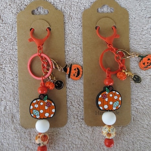 May include: Two orange keychains on brown display cards. Each features a large orange pumpkin charm with white polka dots and a blue patch, a white bead, a patterned bead, and a small orange bead. Additional charms include a pumpkin, a smiley face, and a witch hat.
