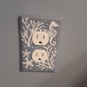 Puede incluir: Una tapa de toma de corriente azul claro con un diseño blanco de caballito de mar, coral y estrella de mar.