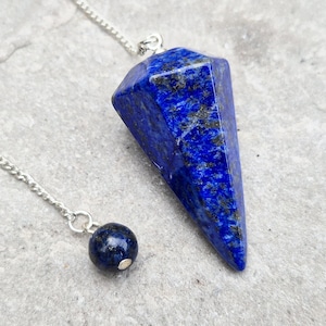 May include: A faceted, deep blue lapis lazuli pendulum with a silver chain and a small, round bead. The gemstone has flecks of gold and sits on a light grey surface. The pendulum is used for divination.