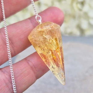May include: A faceted, translucent yellow citrine crystal pendulum suspended from a delicate silver-toned metal chain. The gemstone features a pointed, geometric shape with internal inclusions and a polished surface that catches the light.
