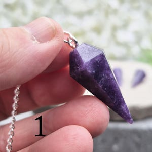 May include: A faceted, deep purple crystal pendulum with a silver chain. The crystal is held in a hand, showcasing its pointed shape and polished surface. The background is blurred, highlighting the crystal's details.