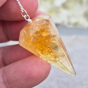 May include: A faceted, cone-shaped pendant made of translucent yellow resin with embedded gold-coloured flakes. The pendant features a silver-toned metal bail at the top, which is attached to a thin silver-coloured chain for suspension.