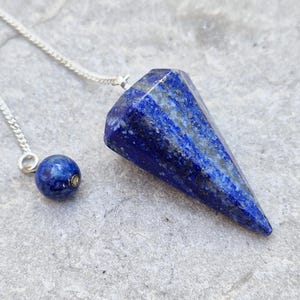 May include: A faceted, deep blue lapis lazuli pendulum with a silver chain and a matching blue sphere. The gemstone pendulum is a cone shape with a silver chain attached to the top. The sphere is attached to the chain.