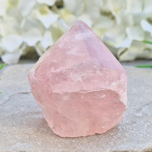 May include: A polished rose quartz crystal point with a pale pink hue and a translucent, multifaceted surface. The stone features a pointed top and a rough, natural base, showcasing internal fractures and variations in its light pink colour.