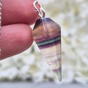 May include: A faceted, cone-shaped fluorite crystal pendant featuring horizontal bands of purple, green, and clear tones. The gemstone is attached to a silver-toned metal chain link at the top, showcasing natural mineral inclusions and variations.