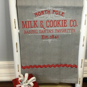 May include: A gray herringbone patterned kitchen towel with red embroidered text that reads "NORTH POLE MILK & COOKIE CO. BAKING SANTA'S FAVORITES Est. 1841". The towel has a red scalloped trim and is displayed on a white ladder.