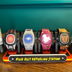 Könnte beinhalten: Eine schwarze und rote "Pixie Dust Refueling Station" hält vier bunte Smartwatches mit verschiedenen Designs, darunter Mickey Mouse und ein Schiff. Zwei FuelRod-Batterien flankieren die Uhren. Die Station trägt den Text "Pixie Dust Refueling Station".