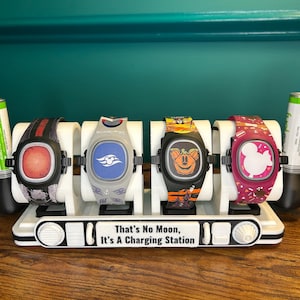 May include: A white charging station holds four smartwatches with various designs, including a red square, a blue cruise ship logo, a Halloween pumpkin, and a pink Mickey Mouse. The base reads "That's No Moon, It's A Charging Station."