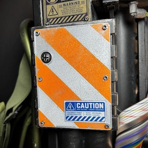 May include: A rectangular metal plate featuring diagonal orange and silver hazard stripes, secured with four corner screws. A blue caution label reads CAUTION HIGH VOLTAGE ENERGY DISCHARGE, DISCONNECT POWER BEFORE SERVICING, OUTPUT 500.000 MHZ, VOLTAGE 1 kilovolts.