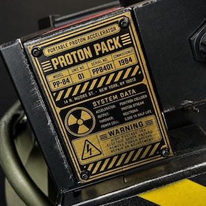 May include: A black proton pack prop featuring a gold-colored metal plate with black text reading Portable Proton Accelerator, Proton Pack, Model PP-84, Unit No. 01, Serial No. PP8401, Commissioned 1984, System Data, and a yellow warning hazard symbol.