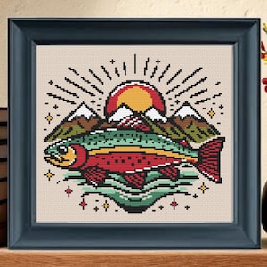 May include: Cross-stitch artwork in a dark blue frame, depicting a fish, mountains, and a sunset. The fish has red, green, and yellow details. The background is a light beige colour. The framed art sits on a wooden surface. The artwork is a square shape.