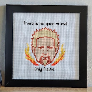 May include: Framed cross-stitch art featuring a stylized face with spiky blonde hair and a goatee, surrounded by flames. The text reads "There is no good or evil, Only Flavor." The artwork is in a black frame.