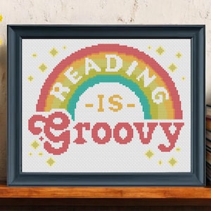 Könnte beinhalten: Ein gerahmtes Kreuzstich-Kunstwerk mit dem Text "READING IS groovy" in einem Regenbogen-Design. Das Kunstwerk befindet sich in einem schwarzen Rahmen und steht auf einem Holzregal. Gelbe Blumen in einer weißen Vase befinden sich links, und ein Stapel Bücher rechts.