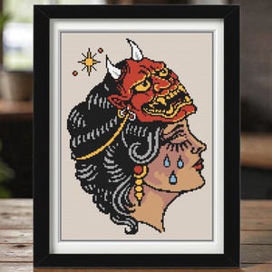 May include: A framed cross-stitch pattern featuring a profile view of a person with dark hair wearing a red Japanese Hannya mask on their head. The design includes blue teardrops on the cheek, a gold earring, and a yellow star symbol in the corner.