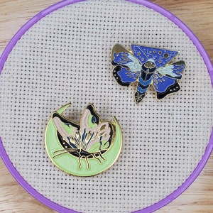 May include: Two enamel pins featuring moth designs. One pin depicts a blue moth with gold accents, the other shows a moth on a green crescent moon. The pins are displayed on a white cross-stitch fabric within a purple embroidery hoop.