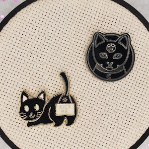 May include: Two black enamel pins featuring a black cat design. One pin shows a cat with a third eye and a crescent moon. The other pin shows a cat with a sign that says "Kiss Me".