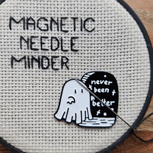 May include: A white ghost-shaped magnetic needle minder with a black tombstone that reads "never been better" on a white canvas background. The text "Magnetic Needle Minder" is embroidered on the canvas.