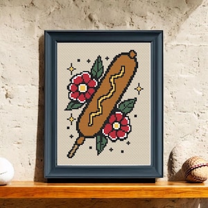 May include: Framed cross-stitch art featuring a corn dog with mustard, surrounded by red flowers and green leaves. The artwork is set against a beige background and framed in a dark blue frame. The style is reminiscent of traditional tattoo art.