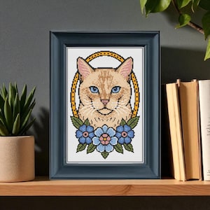 Può includere: Opera d'arte a punto croce incorniciata, raffigurante un gatto con occhi blu e pelo marrone chiaro, circondato da un ovale giallo e fiori blu. La cornice blu scuro è esposta su una mensola di legno.