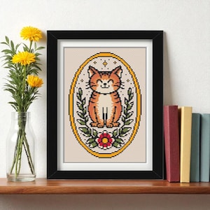 May include: A framed cross-stitch art piece featuring a smiling orange tabby cat within a yellow oval frame. The cat is surrounded by green leaves and a red flower. The artwork is displayed on a wooden shelf with books and a vase of yellow flowers.